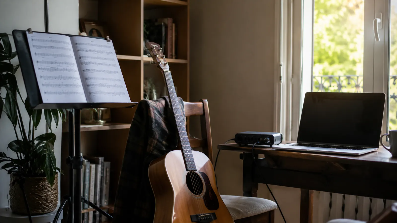 A practice corner by a window with sheet music on a stand, an acoustic guitar leaning on a chair, and a laptop with a compact audio interface on a small table