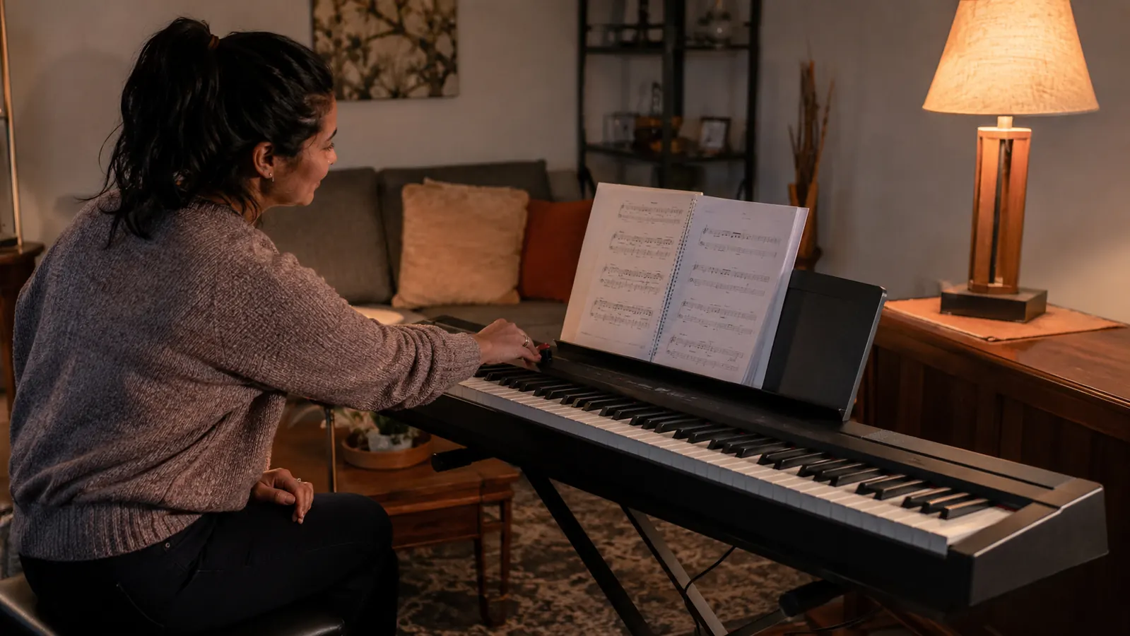 A home practice corner at dusk: an 88-key digital keyboard on a folding stand, sheet music propped on its music rest, and a parent reaching toward its control panel.