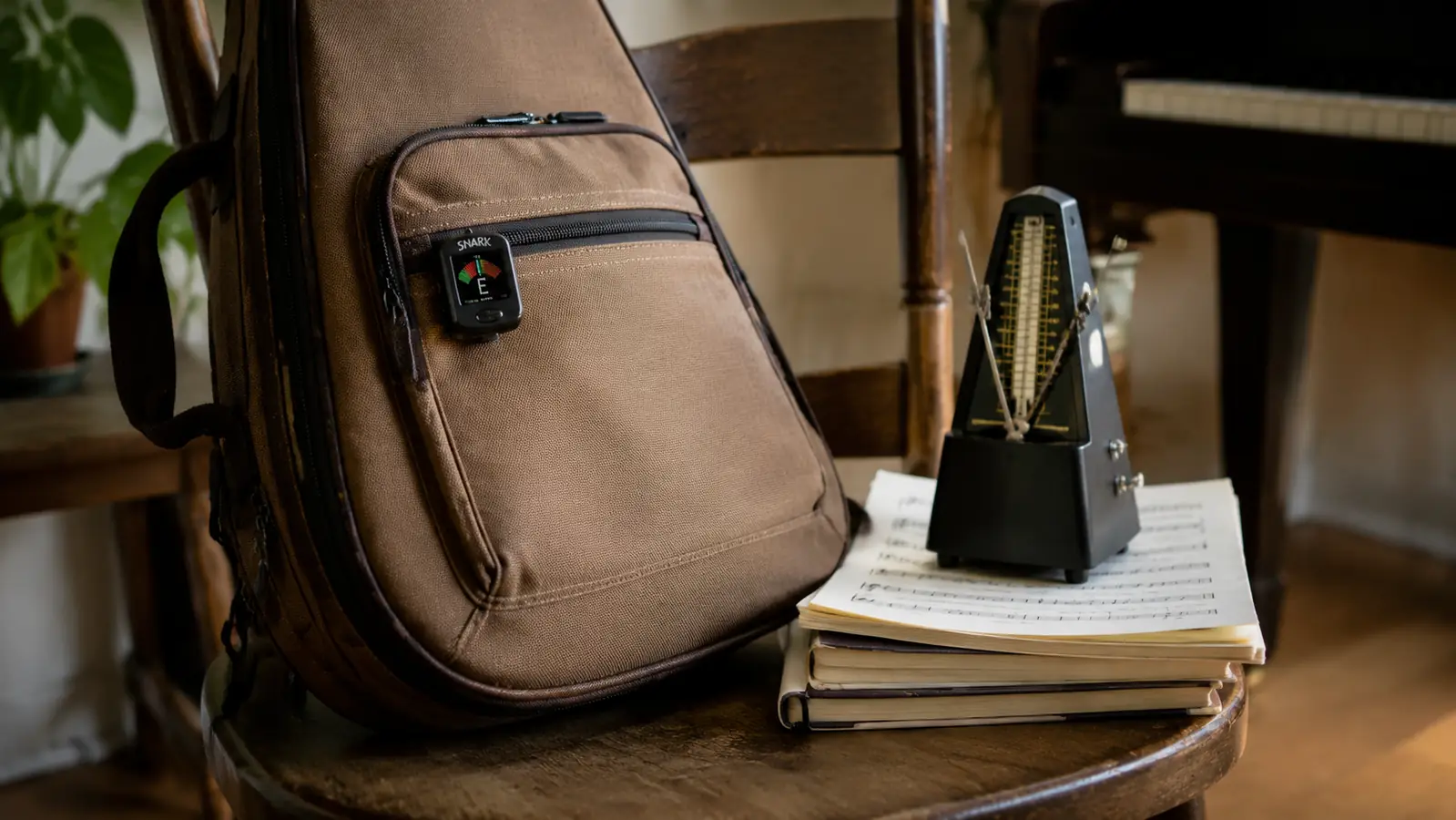 A worn canvas gig bag on a wooden chair with a small clip-on tuner on the outside strap, a metronome and sheet music nearby